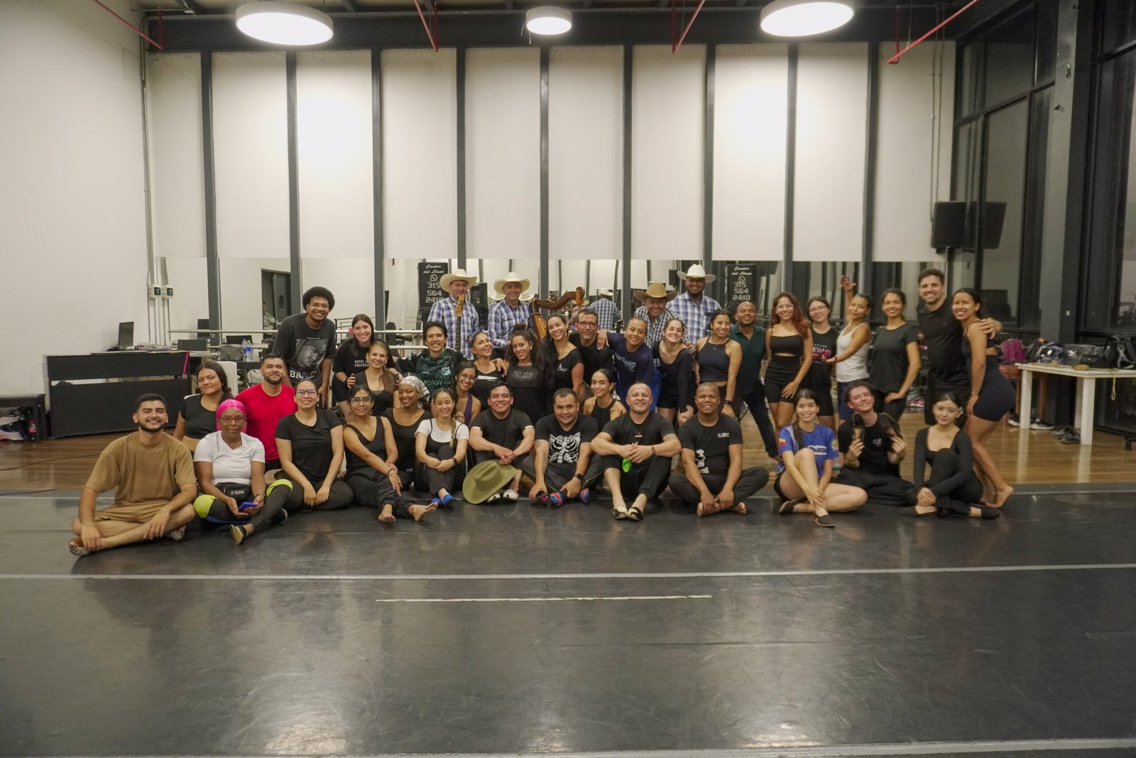 Una fotografía de grupo de más de 30 personas posando en un espacioso estudio de danza. La mayoría de los asistentes visten ropa de entrenamiento negra, mientras que algunos llevan prendas de colores. Al fondo, un grupo de músicos con sombreros de vaquero y camisas a cuadros sostienen instrumentos, incluyendo una arpa. En la pared del fondo, hay pancartas con el texto 'Cántaro del Llano' y números de teléfono. El grupo está distribuido de pie y sentado en el piso reflectante negro.